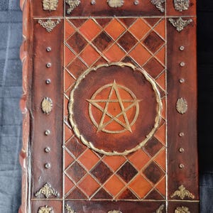 May include: A brown leather-bound journal with a gold pentagram design on the front cover. The cover is decorated with a diamond pattern and gold accents.
