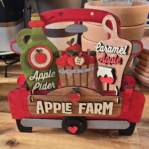 May include: A red wooden truck with a white sign that says "APPLE FARM". The truck is decorated with apples, a bottle of apple cider, and a jar of caramel apple sauce.