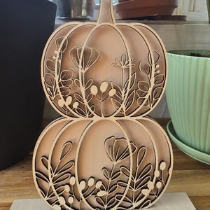 May include: A wooden pumpkin decoration with a layered design. The pumpkin is made of light brown wood and features cut-out floral and leaf patterns. It is a two-tiered design, with each tier resembling a pumpkin shape. The decoration sits on a rectangular wooden base.