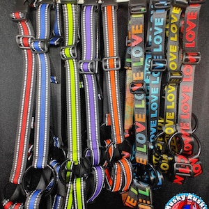 May include: A selection of dog harnesses in a variety of colours, including red, blue, green, purple, and orange. Some harnesses display the word "LOVE" in different colours. Each harness has a black buckle and a metal ring for leash attachment.