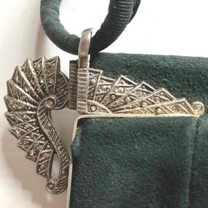 May include: A dark green suede handbag featuring a silver-toned metal clasp adorned with small, sparkling inset stones. The clasp has a fan-like, geometric design and is attached to a dark green fabric handle. The texture of the suede is visible.