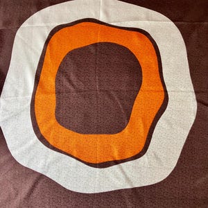 May include: A square scarf featuring a dark brown background with a subtle grid pattern. In the center, a large, irregular white ring surrounds a vibrant orange, amoeba-shaped form. The text Emile sa is visible in the bottom right corner.