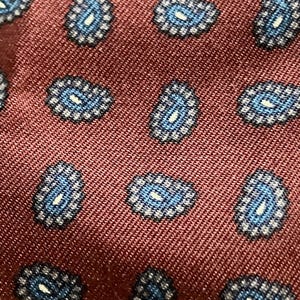 May include: A deep burgundy fabric featuring a repeating pattern of small, intricate paisley motifs. Each paisley shape is rendered in shades of light blue and white, creating a textured, woven appearance across the dark red background surface.