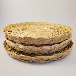 May include: A stack of four woven wicker placemats in a light brown color. The placemats are round and have a braided edge.
