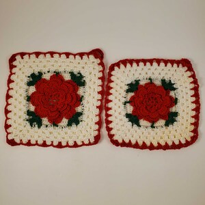 May include: Two crocheted square potholders. Each potholder features a red flower in the center, surrounded by green leaves, on a white background. The edges are trimmed with a scalloped red border. One potholder has a loop for hanging.