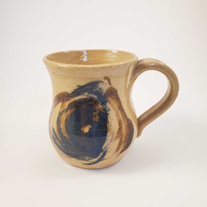 May include: A tan ceramic mug with a blue and brown swirl design. The mug has a rounded body and a handle.