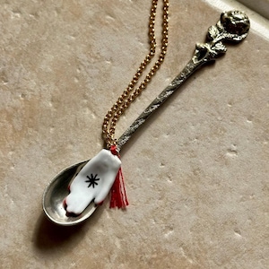 May include: A silver-toned spoon pendant featuring a rose motif on the handle and a small white ceramic hand charm with a black asterisk symbol resting in the bowl. The spoon is suspended from a gold-toned chain with a small red tassel attached.