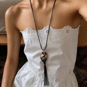 Op de afbeelding: Een zwarte koordketting met een goudkleurige, getextureerde hanger. De hanger is omhuld in een geweven zwart net en heeft een lange zwarte kwast. De ketting wordt gedragen over een witte, strapless top met bloemenborduurwerk.