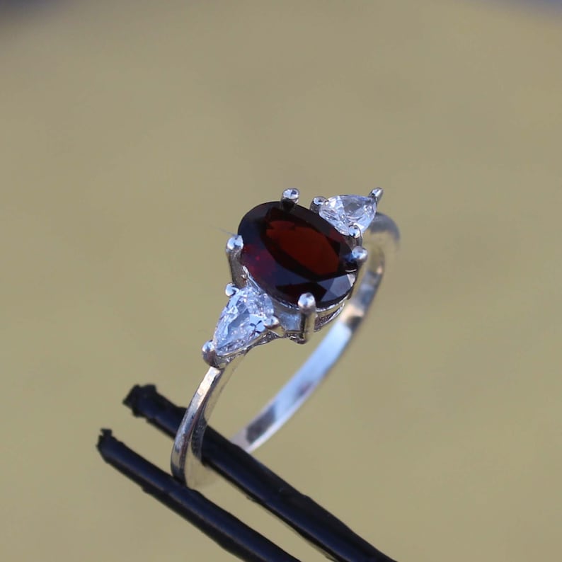 May include: A silver ring with a red oval gemstone and two smaller clear gemstones. The ring is set on a black surface.