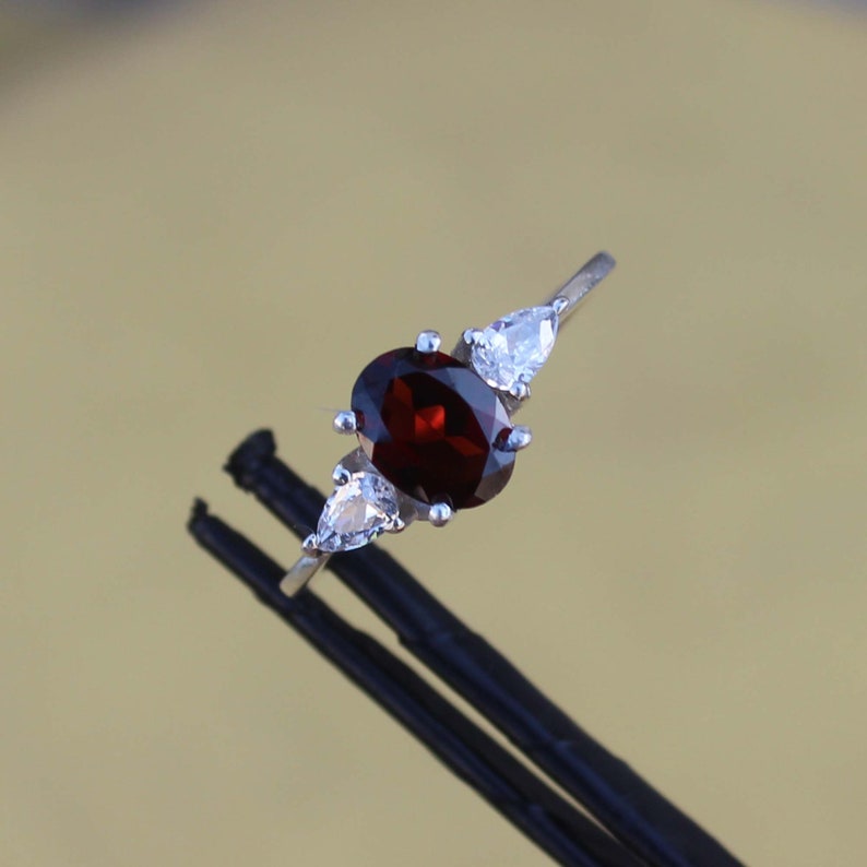 May include: A silver ring with a red oval gemstone and two smaller pear-shaped clear gemstones.