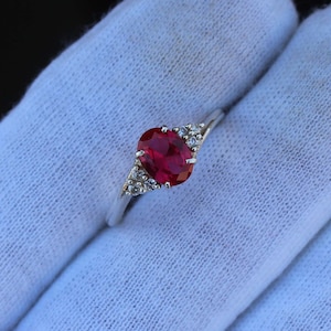 Op de afbeelding: Een zilveren ring met een grote, ovaal geslepen, dieprode edelsteen. Kleine, heldere accentstenen zijn aan weerszijden van de hoofdsteen geplaatst. De ring wordt gepresenteerd op een witte stoffen achtergrond.