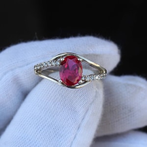 Peut inclure: Une bague en argent ornée d'une grosse pierre rouge foncé de forme ovale. La monture se divise en un double motif de chaque côté de la pierre, avec de petites pierres claires sur le dessus.