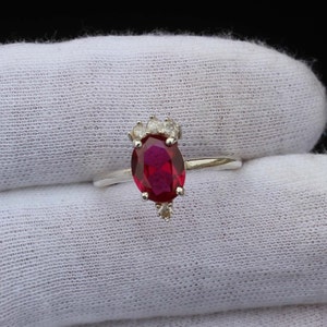Peut inclure: Une bague en argent ornée d'une pierre précieuse ovale rouge foncé. La bague est ornée de petites pierres claires en haut et en bas de la pierre rouge. La bague est posée sur un fond en tissu blanc.