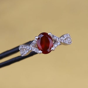 May include: A silver ring with a central oval, deep red gemstone. The band has a twisted design, embellished with small, clear gemstones. The ring is set against a golden-yellow background.