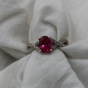 May include: A silver ring with an oval, bright pink gemstone as the centerpiece. The ring's band features an infinity design with small, clear accent stones. The ring is set against a white, textured fabric background.