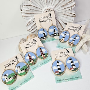 May include: Oval-shaped earrings featuring miniature lighthouse designs. Each earring showcases a unique lighthouse, set against a blue sky with clouds, and framed with a gold-colored border. The earrings are displayed on cards with the text "the daisy butterfly."