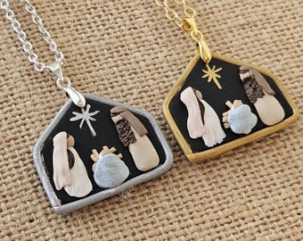 May include: Two necklaces with pendant depicting a nativity scene. One pendant has a silver-colored frame and chain, the other has a gold-colored frame and chain. The pendants feature a black background with figures in beige and blue.