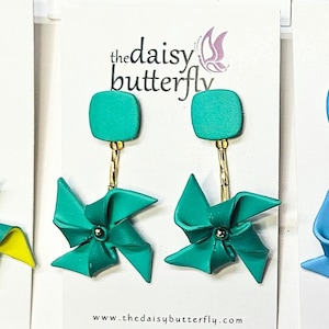 May include: Teal blue square dangle earrings with a teal blue windmill charm. The earrings are on a white card with the text "the daisy butterfly" and "www.thedaisybutterfly.com".