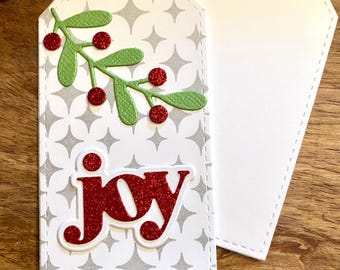 May include: A white gift tag with a gray and white geometric pattern. The tag has a red glitter word "joy" and a green holly sprig with red berries. The tag has a hole punched in the top for string.