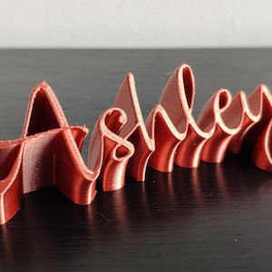 May include: A 3D printed nameplate with the name "Ashley" in a cursive font. The nameplate is made of a red plastic material.