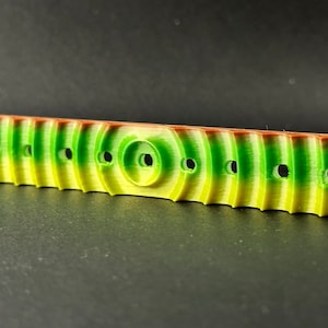 May include: A green, yellow, and brown plastic 3D printed object with a series of holes and a rounded center section.