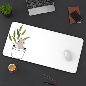 May include: White desk mat with a cartoon cat sitting in a pot of green plants. The mat is on a black surface with a laptop, a notebook, and a computer mouse.