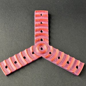 Puede incluir: Un objeto de plástico rosa y naranja de tres puntas con un centro circular y varios agujeros. El objeto tiene una superficie texturizada y parece estar impreso en 3D.