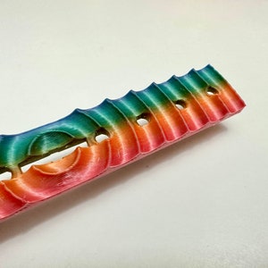 May include: A rainbow-colored plastic object with a wavy design and multiple holes. The object is about 10 inches long.