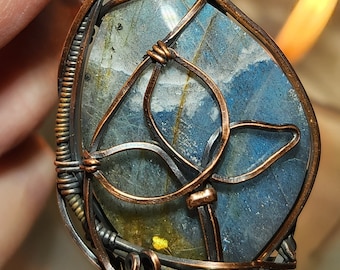 Peut inclure: Pendentif artisanal avec une pierre précieuse bleue et grise, finement enveloppée de fil de cuivre et d'argent. Le fil forme un motif décoratif autour de la pierre, avec un petit accent de coquillage blanc en haut. Un bijou unique.