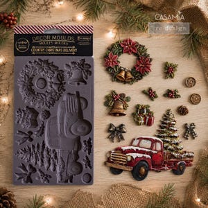 May include: A grey silicone decor mould labelled Country Christmas Delivery featuring cavities for a wreath, bells, a truck, a tree, and bows. Beside it are finished decorative pieces including a poinsettia wreath, bells, gifts, and a red truck with a tree.