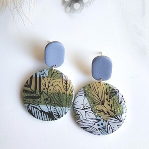 Op de afbeelding: Een paar oorhangers met een rond, gepatroneerd ontwerp met groene, gouden en zwarte accenten. De oorhangers hebben een lichtblauwe, ovale bovenkant.