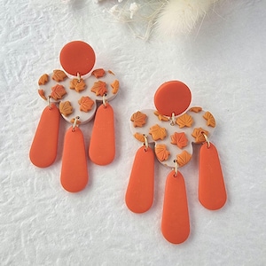 May include: A pair of orange and white dangle earrings with a leaf pattern. The earrings have three orange teardrop-shaped dangles on each side.