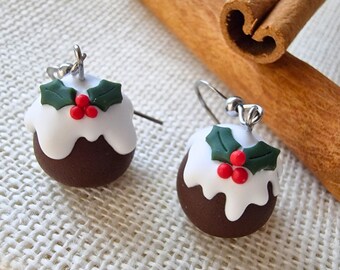 May include: A pair of Christmas pudding earrings. Each earring features a dark brown sphere topped with white icing, green holly leaves, and red berries. The earrings hang from silver-colored hooks.