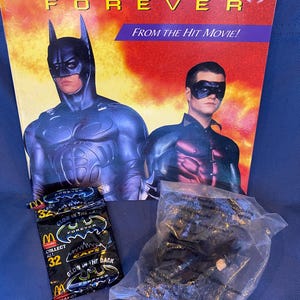 May include: A Batman Forever book with Batman and Robin on the cover, along with a McDonald's collectible and a Batman toy in a plastic bag. The book has the text "FROM THE HIT MOVIE!" and "Golden Books". The McDonald's collectible says "GLOW IN THE DARK".