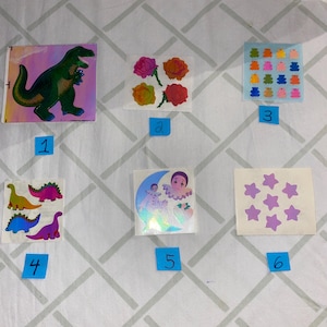 May include: A set of ten vintage holographic stickers featuring various designs, including dinosaurs, flowers, a clown, stars, and frogs. The stickers are arranged in a grid on a white surface.