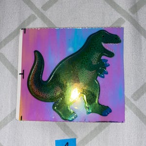 May include: A green holographic sticker of a dinosaur on a pink, blue, and purple iridescent background.