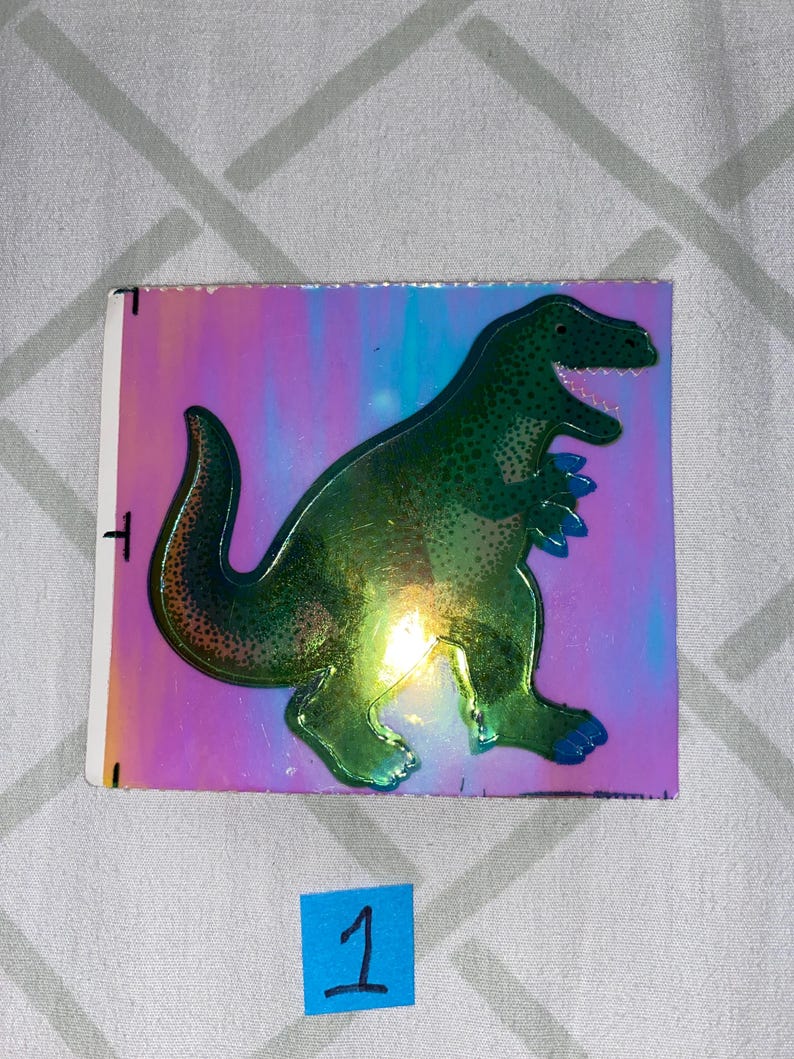 May include: A green holographic sticker of a dinosaur on a pink, blue, and purple iridescent background.