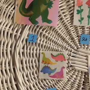 May include: A white wicker basket with four stickers on it. The stickers are of dinosaurs. One sticker is of a green dinosaur, another is of a yellow dinosaur, another is of a pink dinosaur, and the last is of a blue dinosaur. The basket has numbers 1, 2, 3, and 4 written on blue sticky notes.