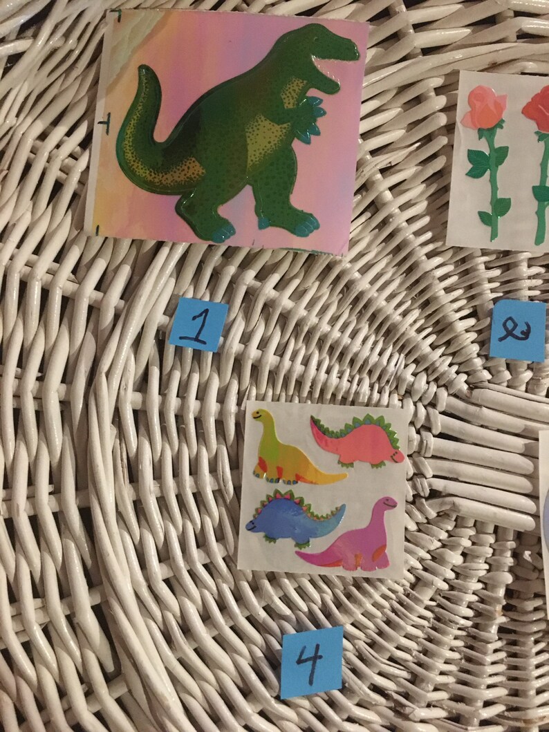 May include: A white wicker basket with four stickers on it. The stickers are of dinosaurs. One sticker is of a green dinosaur, another is of a yellow dinosaur, another is of a pink dinosaur, and the last is of a blue dinosaur. The basket has numbers 1, 2, 3, and 4 written on blue sticky notes.