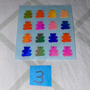 May include: A sheet of 16 iridescent holographic bear stickers in various colors, including green, orange, pink, blue, and red. The sheet is on a light blue background. The number 3 is written on a blue sticky note below the sheet.