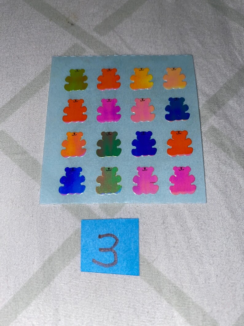May include: A sheet of 16 iridescent holographic bear stickers in various colors, including green, orange, pink, blue, and red. The sheet is on a light blue background. The number 3 is written on a blue sticky note below the sheet.