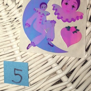 May include: A vintage sticker sheet with three holographic stickers. The stickers feature a purple and blue clown on a crescent moon, a purple and pink clown with a ruffled collar, and a pink heart with a pink flower.