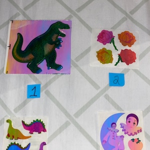 May include: A sheet of holographic stickers featuring a green dinosaur, three pink, yellow, and red roses, and a purple clown on a crescent moon.
