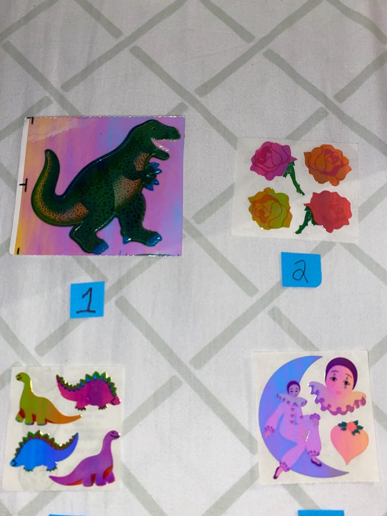 May include: A sheet of holographic stickers featuring a green dinosaur, three pink, yellow, and red roses, and a purple clown on a crescent moon.