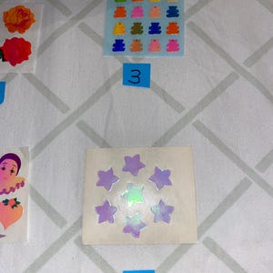 May include: A sheet of colorful gummy bear stickers with a blue background. There are 16 stickers in total, each with a different color. The sheet is labeled with the number "3". Below the sheet is a sheet of iridescent star stickers with a white background. There are 8 stickers in total, each with a different shade of purple and green. The sheet is labeled with the number "6".