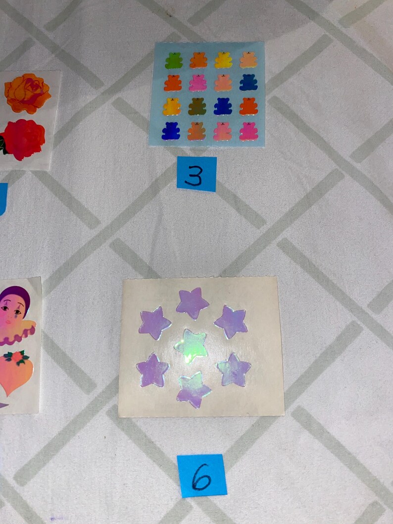 May include: A sheet of colorful gummy bear stickers with a blue background. There are 16 stickers in total, each with a different color. The sheet is labeled with the number "3". Below the sheet is a sheet of iridescent star stickers with a white background. There are 8 stickers in total, each with a different shade of purple and green. The sheet is labeled with the number "6".