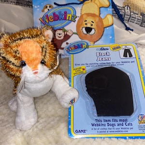 May include: A plush tiger toy with orange and brown stripes, a Webkinz gift bag, and a package of black jeans for Webkinz pets. The gift bag features cartoon animals. The jeans package reads "Black Jeans" and "This item fits most Webkinz Dogs and Cats."