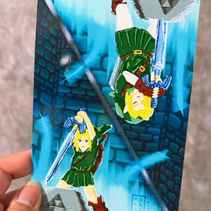 May include: A rectangular art print featuring a mirrored illustration of a blonde character in a green tunic holding a blue sword. The composition is divided diagonally, showing the character upright and inverted against a blue stone wall background.
