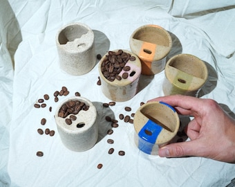 May include: Five small ceramic cups with a divided section, four are white with speckles, one is white with blue and beige stripes. One cup is filled with coffee beans.