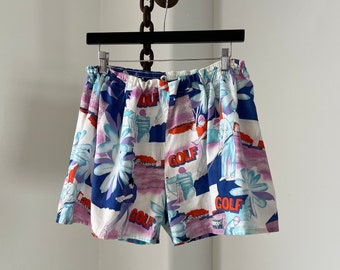 May include: White cotton shorts with a colourful print of golfers and the word "GOLF" repeated throughout. The shorts have a drawstring waist.